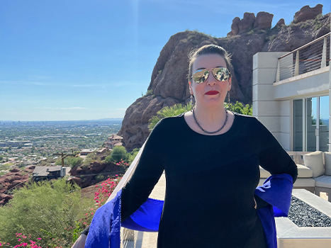 Monica in black dress and purple sash standing on balcony in sunglasses